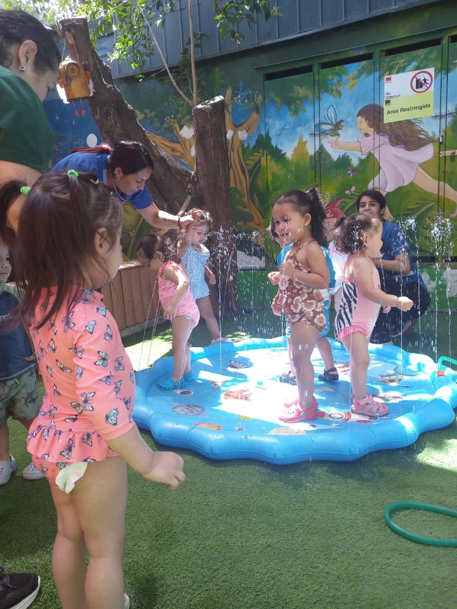 Niñas del jardín juegan con agua en una piscina inflable en el patio, frente a un mural de bosque